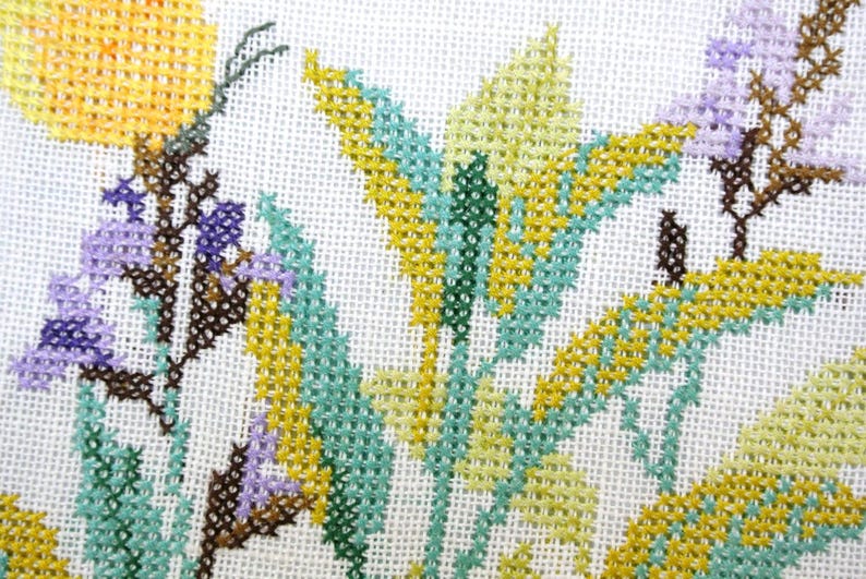 Puede incluir: Primer plano de un dise&ntilde;o floral de punto de cruz con flores amarillas, verde claro y moradas con tallos y hojas marrones. La labor de aguja est&aacute; sobre un fondo de tela blanca.