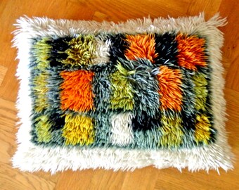 May include: A rectangular throw pillow featuring a shaggy, high-pile texture. The center panel displays a grid of multicolored yarn squares in shades of orange, olive green, dark gray, and yellow, surrounded by a thick, fluffy white border.