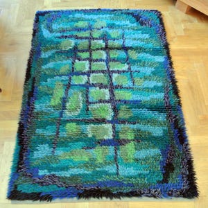 May include: A rectangular rug with a geometric pattern in shades of blue, green, and black. The rug has a shaggy texture and a dark border. The design features squares and lines, creating a modern, abstract look. The rug is placed on a wooden floor.