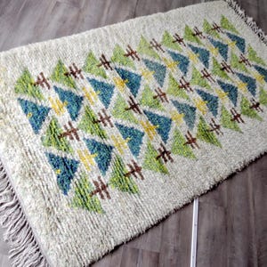 Peut inclure: Un tapis rectangulaire à poils longs blanc cassé orné d'un motif géométrique répétitif de triangles dans des tons vert lime, bleu sarcelle et jaune, agrémenté de petits détails en croix marron et de longues franges aux deux extrémités.