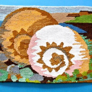 May include: Woven wall hanging depicting two large, stylised seashells in shades of tan, white, and pink. The background shows a coastal scene with blue water, green foliage, and brown cliffs. The artwork is on a blue background.