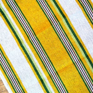 May include: A woven textile with a striped pattern. The design features alternating bands of yellow, white, and green, with thin black and red stripes. The textile is likely a table runner or decorative cloth.