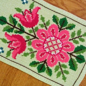 May include: A green and white embroidered runner with pink flowers. The runner features a floral design with three large pink flowers and green leaves.