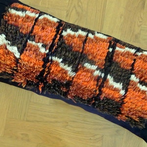 May include: A long, rectangular pillow with a black fabric backing and a colorful, textured front. The front is made of a thick, woven fabric with a geometric pattern in shades of orange, brown, and white.