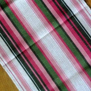 Puede incluir: Una tela de tejido blanco, rosa, verde y negro con textura tejida. La tela está doblada por la mitad y las rayas corren en diagonal.