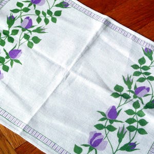 Puede incluir: Paño rectangular blanco con un diseño floral. El diseño presenta rosas moradas y hojas verdes dispuestas a lo largo de los bordes. Un fino borde morado enmarca el paño. El paño está colocado sobre una superficie de madera.