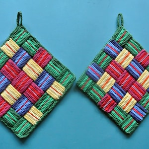 Pode incluir: Dois pegadores de panela de crochê coloridos com um padrão tecido em verde, amarelo, vermelho, azul e laranja. Os pegadores têm um laço para pendurar.
