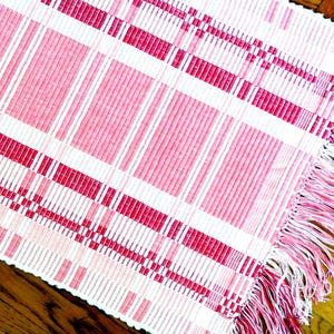 May include: A woven pink and white patterned textile with a fringed edge. The design features a grid of vertical and horizontal stripes in varying shades of pink and white. The fringe is a lighter shade of pink.