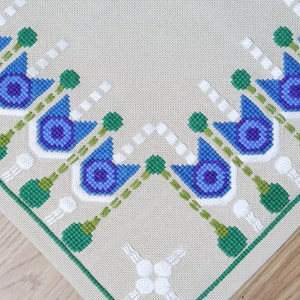 Puede incluir: Una bordadura de punto de cruz beige con un borde verde y un patrón de flores azules y blancas. Las flores tienen un centro morado y están dispuestas en un diseño simétrico.