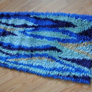 Puede incluir: Una alfombra de pelo azul y beige con un patrón ondulado y flecos en todos los lados.