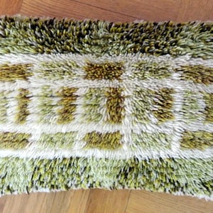 Peut inclure: Coussin rectangulaire décoratif avec une surface touffue et texturée. Le coussin présente un motif de formes rectangulaires vertes, blanches et marron. Le coussin est posé sur un plancher en bois.