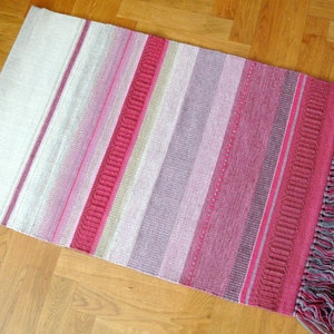Puede incluir: Una alfombra de rayas rosas y blancas con un borde de flecos. La alfombra tiene un sutil efecto degradado, con las rayas rosas que se desvanecen gradualmente a blanco.