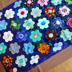 May include: A handmade quilt featuring a vibrant array of colorful, hexagonal flower shapes. The flowers are made from various patterned fabrics, including floral, striped, and polka dot designs, set against a dark blue background with a brown border.