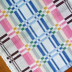 May include: A white woven placemat with a colorful plaid pattern in shades of blue, pink, yellow, and green. The placemat has a white crocheted border.