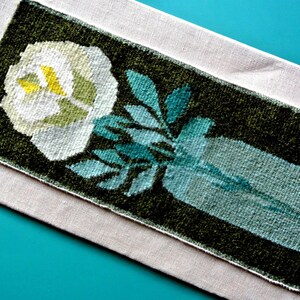 May include: A rectangular, woven floral design featuring a white flower with yellow center and teal leaves in a vase. The design is set against a dark green background and rests on a light beige fabric.