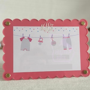 Op de afbeelding: Een roze, geschulpte fotolijst met gouden accenten. De lijst bevat een babythema-ontwerp met babykleding en een fopspeen. Een houten hondenspeelgoed en een kralenketting zijn op de achtergrond.