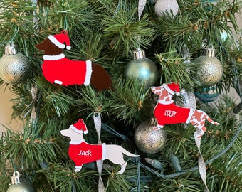 May include: Three dachshund dog ornaments in red and white Santa suits with the names Jack, Colby, and a third name that is not visible hanging on a Christmas tree.