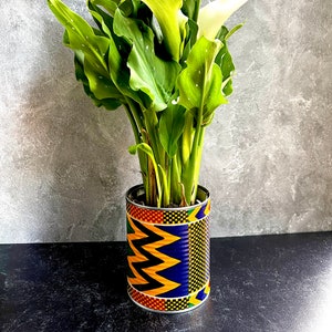 May include: A potted calla lily plant in a decorative tin can. The can is wrapped with a colorful African print featuring yellow zigzags, blue and red geometric patterns. The plant has large green leaves and white flowers.