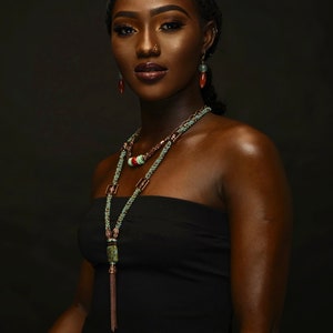 May include: A woman wearing a black strapless top and elaborate beaded jewelry. The necklace features turquoise, red, and green beads with a copper-colored chain and a rectangular pendant. Matching earrings complete the look.