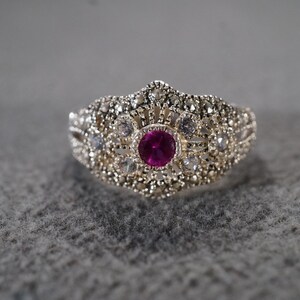 May include: Silver-toned ring with a central, round, deep pink gemstone. The ring features a detailed, ornate design with smaller clear and white stones surrounding the central gem. The ring's band has a textured appearance.