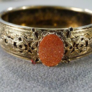 May include: A gold-toned bangle bracelet with an intricate floral design and a large, oval-shaped, orange sunstone.