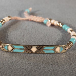 May include: A turquoise and white beaded bracelet with a gold metal charm featuring a geometric design. The bracelet is tied with a pink cord.