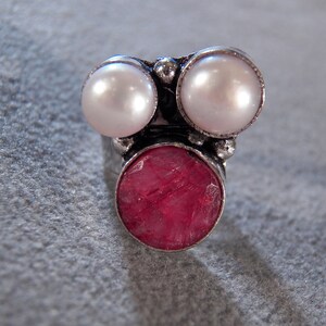 May include: A silver ring with three round stones. The center stone is a large, deep red, and the two smaller stones on either side are white.