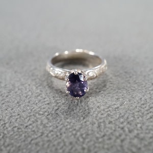 May include: A silver ring with an oval, dark purple gemstone. The ring band has a detailed floral pattern. The gemstone is set in a prong setting, and the ring is displayed on a gray surface.