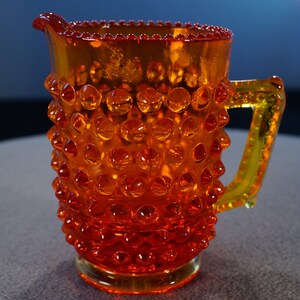 May include: A small orange glass pitcher with a textured, bumpy surface and a handle. The pitcher has a rounded body and a flared lip.