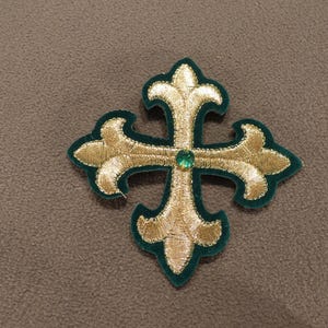 May include: A decorative cross-shaped patch featuring metallic gold embroidery in a fleur-de-lis style. The cross is outlined with a dark green felt border and features a single, small, round, faceted green gemstone set at the center point.