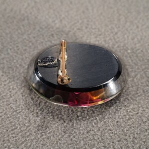 K&ouml;nnte beinhalten: Die R&uuml;ckseite einer Brosche mit einem schwarzen ovalen Cabochon-Stein, der in eine silberfarbene Metallfassung gefasst ist. Die Brosche hat einen goldfarbenen Stift und eine Schlie&szlig;e.