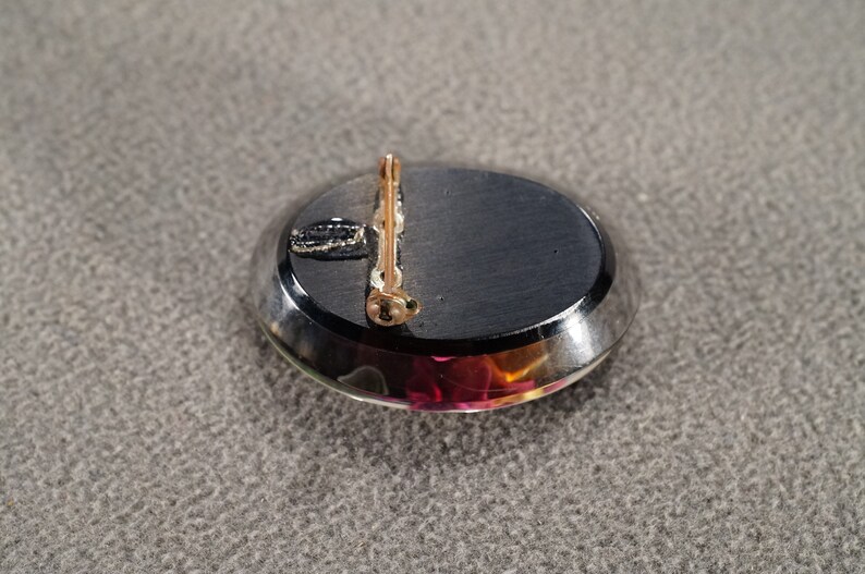 K&ouml;nnte beinhalten: Die R&uuml;ckseite einer Brosche mit einem schwarzen ovalen Cabochon-Stein, der in eine silberfarbene Metallfassung gefasst ist. Die Brosche hat einen goldfarbenen Stift und eine Schlie&szlig;e.