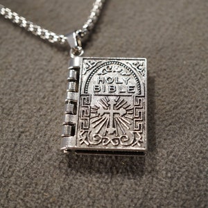 May include: A silver chain necklace with a silver pendant shaped like a closed book. The book has an inscription that reads "HOLY BIBLE" and a cross with rays of light emanating from it.