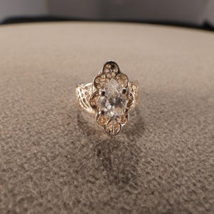 May include: A silver-toned ring with a pear-shaped, clear gemstone at the center. The ring features an ornate filigree design around the gemstone and along the band, creating a vintage aesthetic. The ring is set on a neutral-toned surface.