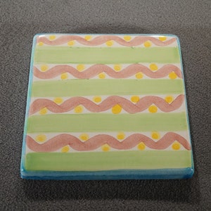 May include: A square ceramic tile with a green and pink striped pattern. The stripes are decorated with a wavy line and yellow dots.