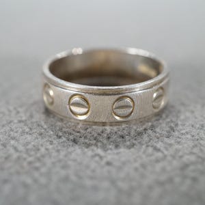 May include: A silver ring with a textured band and evenly spaced, circular screw head designs. The ring is displayed on a gray surface, showcasing its polished metal finish and detailed craftsmanship. A classic piece of jewelry.