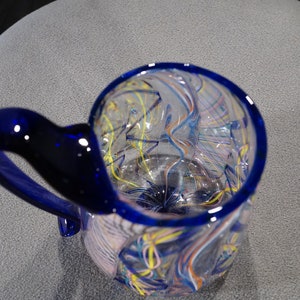 May include: A blue glass mug with a swirling pattern of white, yellow, and blue lines. The mug has a blue handle.