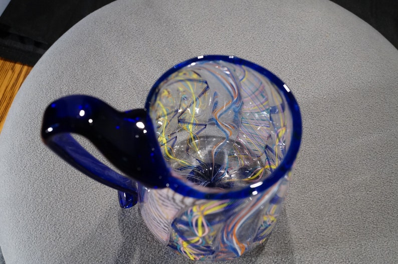 May include: A blue glass mug with a swirling pattern of white, yellow, and blue lines. The mug has a blue handle.