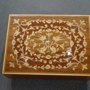 May include: Rectangular wooden box with a decorative inlaid design. The box features a light brown border and a dark brown top with an intricate floral and bird pattern in cream-colored inlay. The box is likely used for jewelry or small keepsakes.