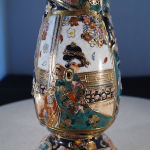 May include: A ceramic vase with a gold and green floral design and a depiction of a woman in a kimono. The vase has a round base and a narrow neck.