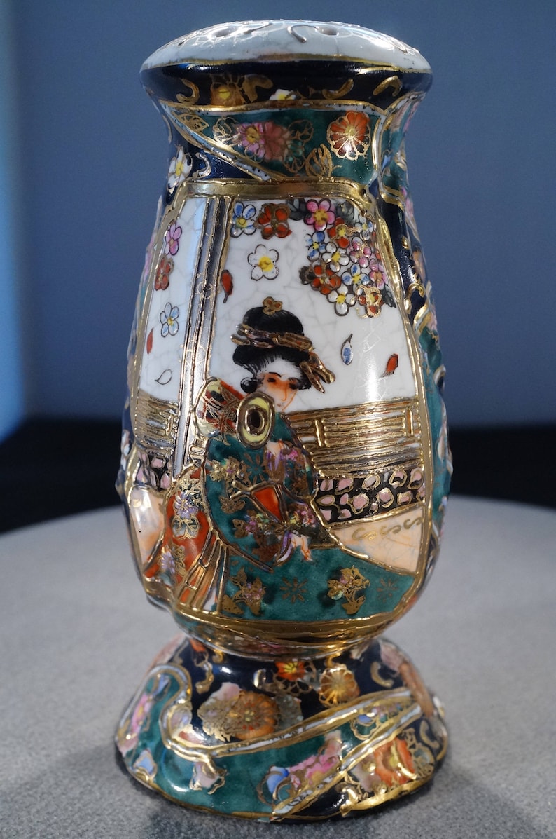 May include: A ceramic vase with a gold and green floral design and a depiction of a woman in a kimono. The vase has a round base and a narrow neck.
