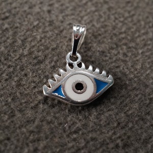 May include: A silver charm in the shape of a blue evil eye with a white center and a black pupil. The charm has a loop at the top for attaching to a necklace or bracelet.