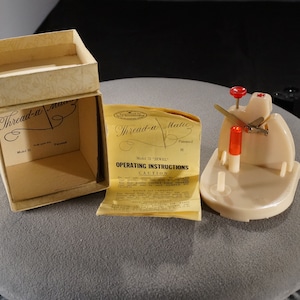 May include: A vintage white plastic Thread-a-Matic threader with red knobs and a metal needle. The threader is in its original box with instructions. The box is beige with the words "Thread-a-Matic" printed on it.