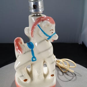 May include: A white ceramic lamp shaped like a rocking horse with blue and pink accents. The lamp has a white cord and a metal socket.
