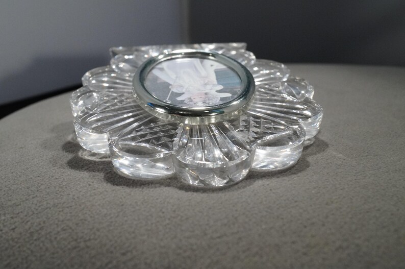 May include: A clear glass picture frame with a flower-shaped design. The frame has a silver metal rim around the photo opening.