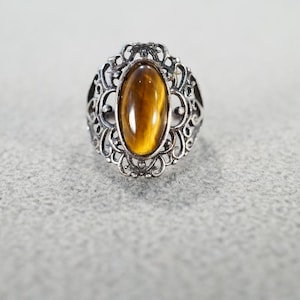 May include: A silver-toned ring with an oval, amber-colored stone. The ring features an ornate, filigree design around the stone. The stone has a striped pattern, and the ring is set on a light gray surface.