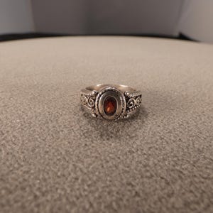 May include: Silver-toned ring with an oval, reddish-brown gemstone at the center. The ring features ornate scrollwork and detailing around the gemstone and on the band. The ring is set against a neutral background.