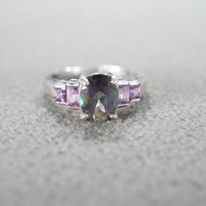 May include: A silver ring featuring an oval, iridescent center stone flanked by two smaller, rectangular purple stones on each side. The ring is set against a neutral gray background.