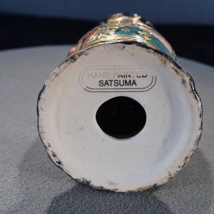 May include: The base of a hand-painted Satsuma ceramic vase featuring a gold and green floral design. The vase is marked "Hand Painted Satsuma" on the underside.
