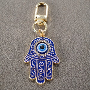 May include: A gold-colored keychain with a blue hamsa hand charm. The hamsa hand has a blue evil eye in the center and is decorated with gold swirls and rhinestones.